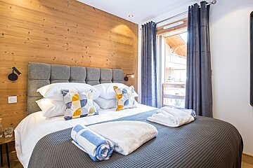 Cozy chalet-style bedroom with a wooden paneled wall, comfortable bed, and balcony window framed by dark curtains.