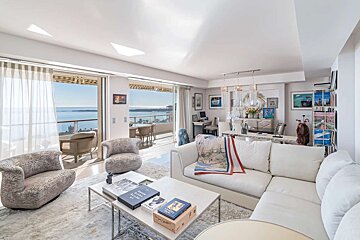 Bright living room with white furniture, patterned chairs, and art, opening to a balcony with stunning ocean views.