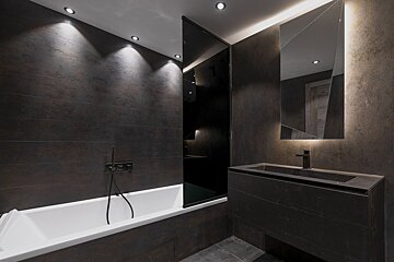 A modern, dark-tiled bathroom features a white tub, dark vanity with a backlit mirror, and recessed ceiling lights for a sleek, moody ambiance.