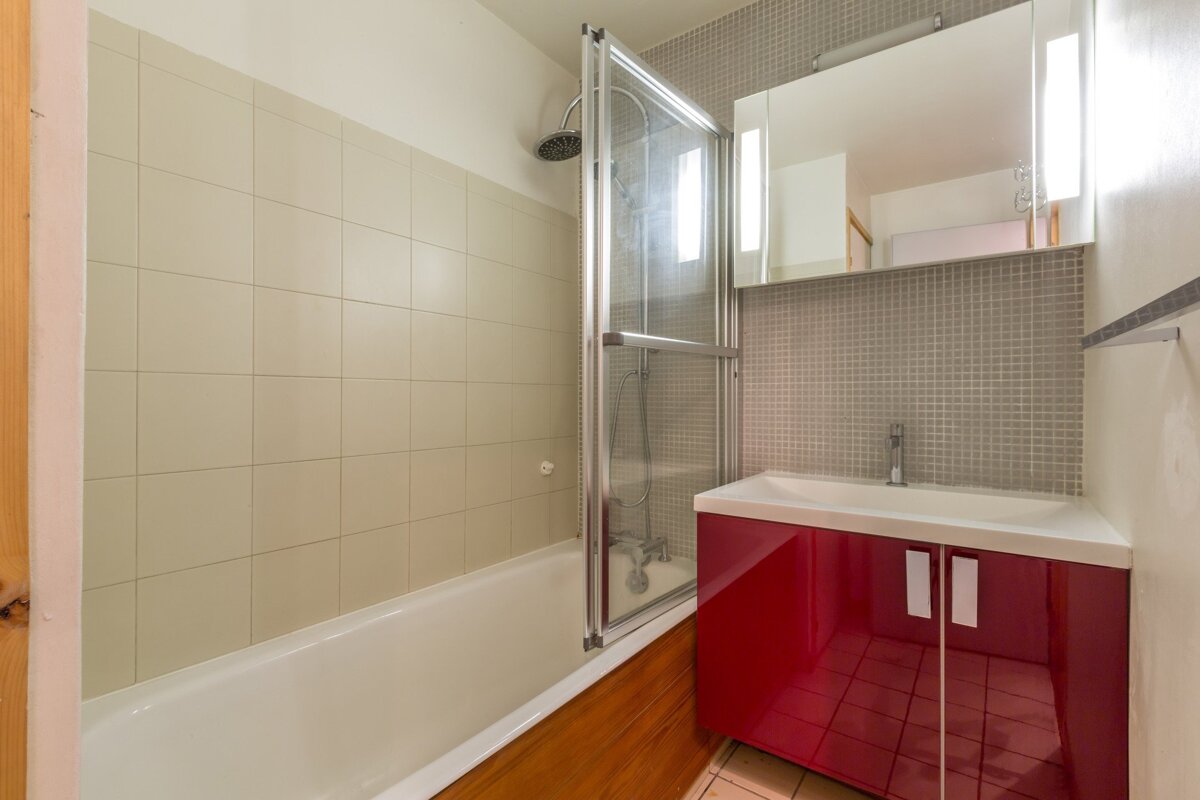 A compact bathroom featuring a bathtub with a glass screen, beige tiles, and a vibrant red sink vanity next to a mirrored cabinet.