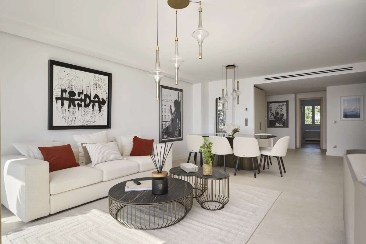 A bright, modern open-plan living and dining room with a white sofa, abstract art, nested black coffee tables, and a dining area with pendant lights.