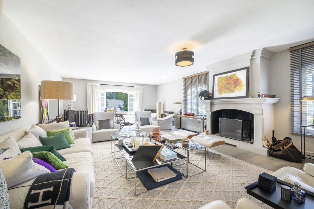 A bright, spacious living room featuring white sofas with colorful pillows, modern glass coffee tables, a large fireplace, and outdoor access.