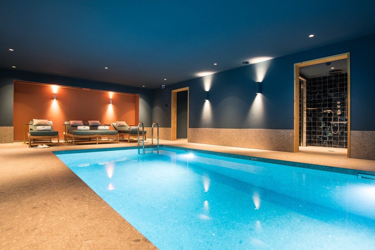 A modern, chic indoor swimming pool with deep blue walls, a warm orange accent, lounge chairs, and a tiled shower area.