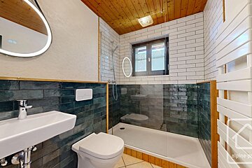 Modern bathroom with wooden ceiling, dark green & white subway tiles. Features a sink, toilet, glass shower, illuminated mirror, and towel warmer.