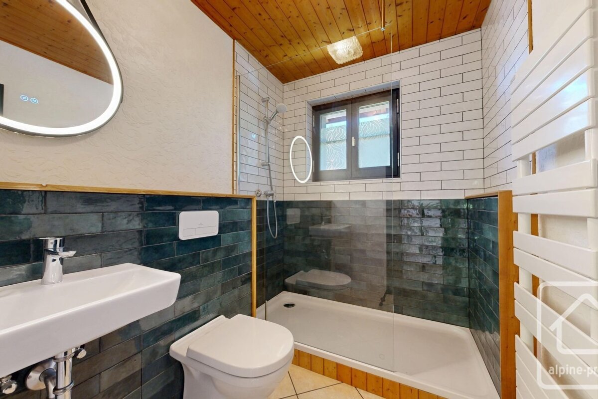 A modern bathroom featuring blue-green tiles, a glass shower, a white sink, a wood ceiling, and a backlit mirror.