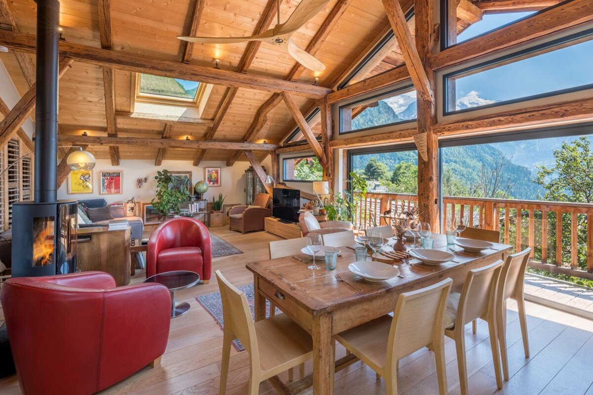 A cozy, rustic chalet interior with exposed wood beams, a fireplace, and panoramic mountain views through large windows. Open-plan living and dining space.