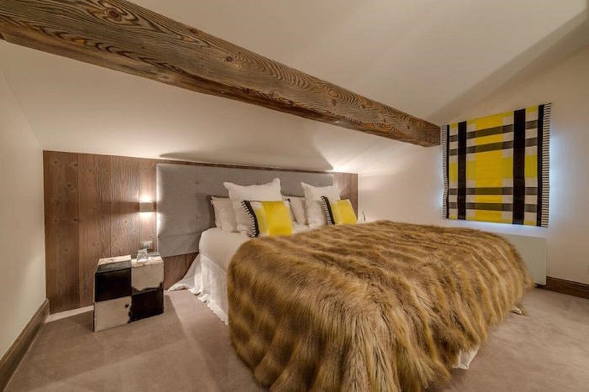 A cozy bedroom features a large bed with a brown fur blanket, yellow accent pillows, a prominent wooden ceiling beam, and wood paneling.