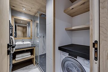 A modern bathroom with wood accents features a vanity, shower, and a hidden laundry area with a washing machine and shelves behind a partially open door.