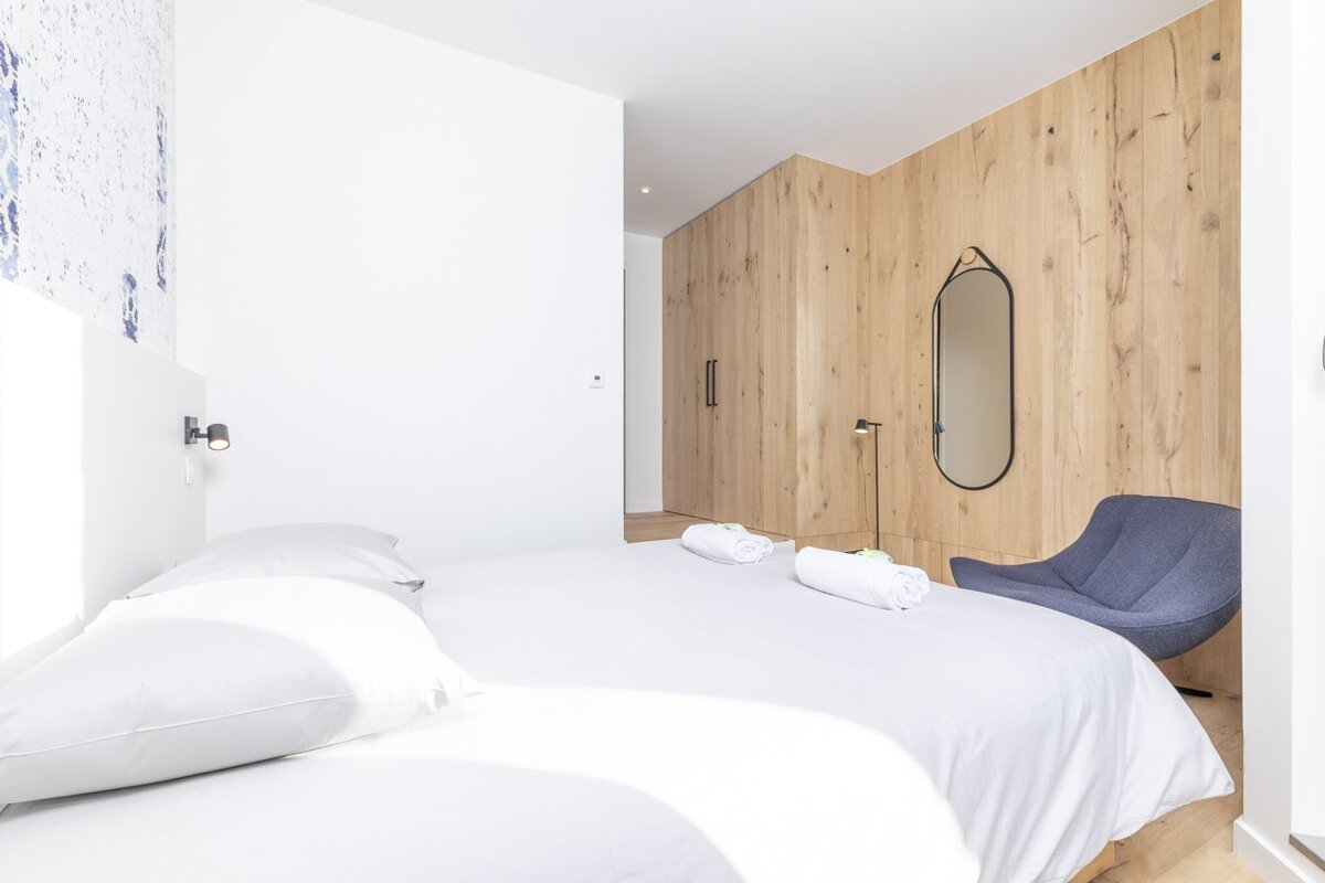 A bright, contemporary bedroom featuring a white bed with rolled towels, a light wood paneled wall with a black mirror, and a blue accent chair.