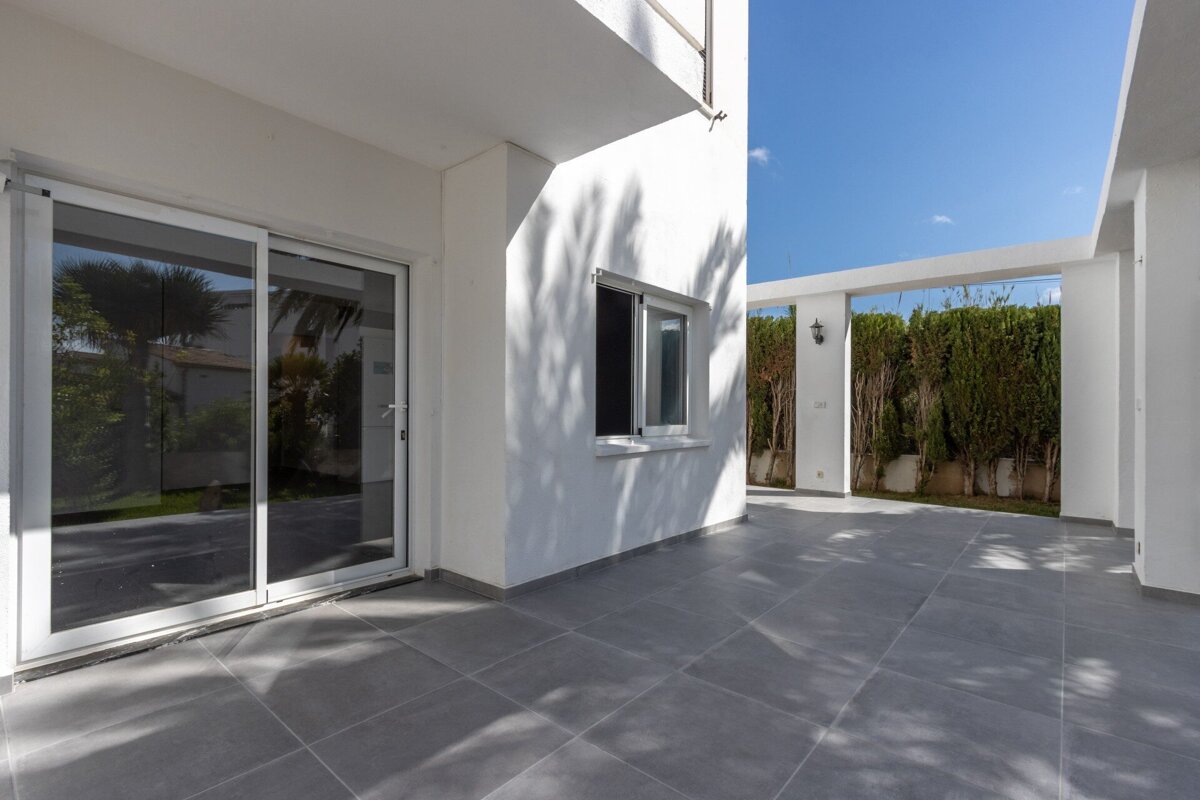 A bright outdoor patio with grey tiled floor, white building, sliding glass doors, a window, and a hedgerow under a blue sky with tree shadows.
