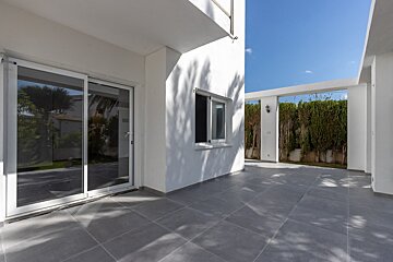 A bright outdoor patio with grey tiled floor, white building, sliding glass doors, a window, and a hedgerow under a blue sky with tree shadows.
