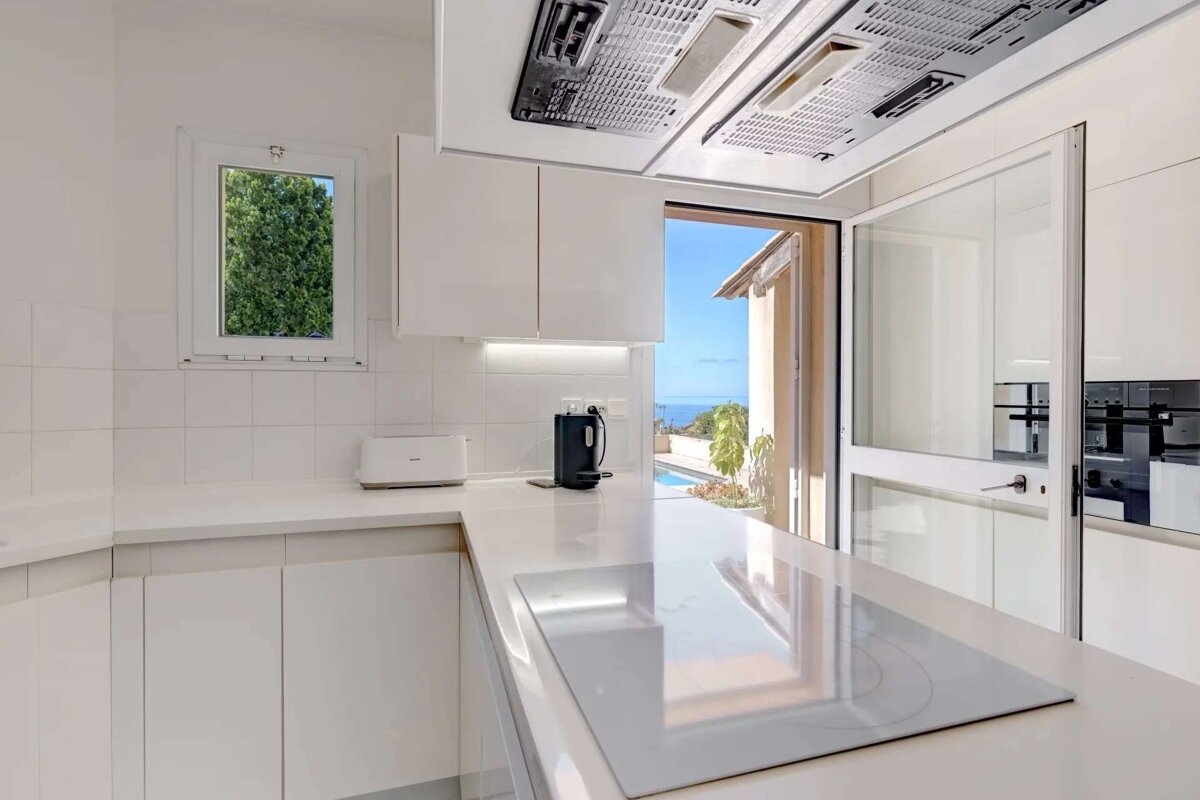 A bright, modern white kitchen with sleek appliances, an induction hob, toaster, and kettle. It opens to a stunning ocean view patio with a pool.