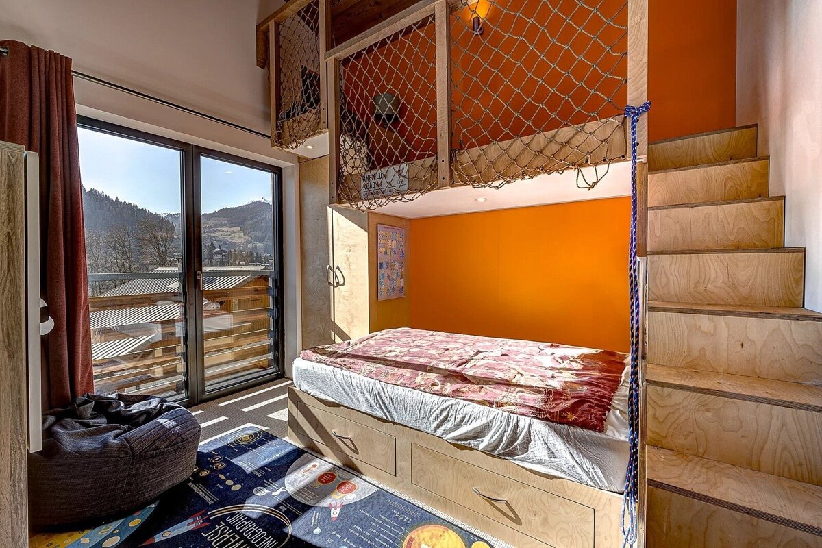 A vibrant kids' room featuring net-enclosed loft beds, an orange accent wall, a bed with storage, and a large window overlooking mountains.