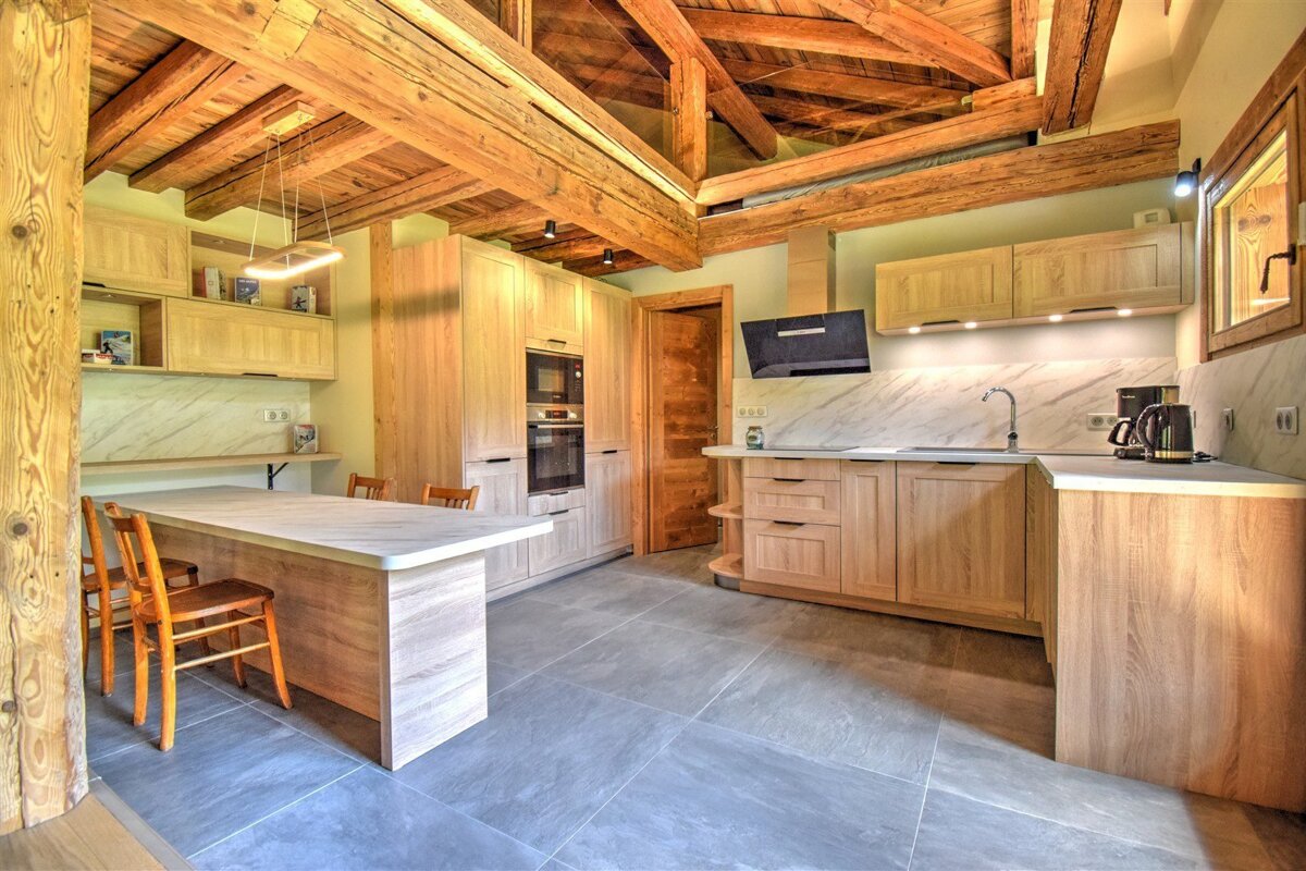 A rustic-modern kitchen with light wood cabinetry, exposed wooden ceiling beams, a large island with seating, and gray tiled flooring.