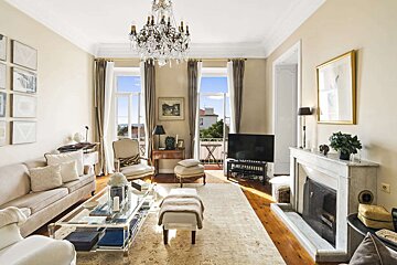 A bright, classic living room featuring a crystal chandelier, large windows to a balcony, a fireplace, and refined furniture in neutral tones.