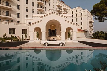 A white vintage convertible is parked in front of a grand white hotel with arched entranceways, reflected in a still pool.