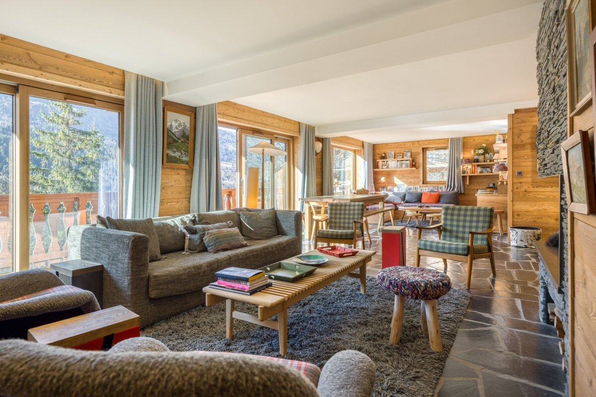 A cozy, sunlit chalet living room with wood-paneled walls, comfortable seating, a stone fireplace, and large windows revealing mountain views.