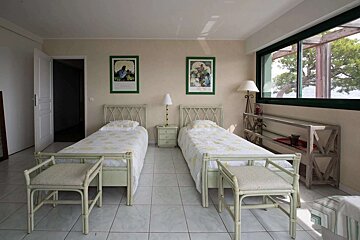 A cozy bedroom features two twin beds with light bedding, rattan-style furniture, framed art, and a large window with an outdoor view.