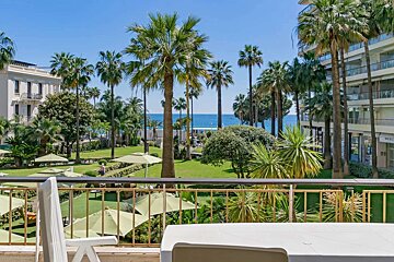 Sunny balcony view of a lush green garden with numerous palm trees, leading to a distant blue ocean, flanked by buildings under a clear sky.