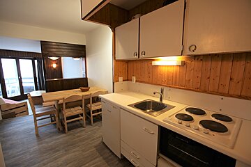 A cozy apartment interior featuring a compact kitchen with wood paneling, a wooden dining table, and a living area with large glass doors leading outside.