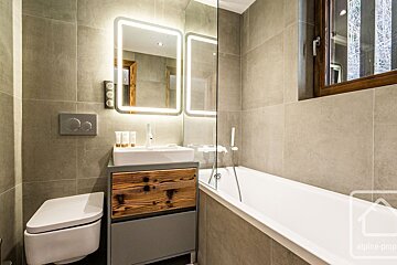 A modern bathroom features beige/grey tiles, a white bathtub, a sleek wooden vanity with a sink and illuminated mirror, and a wall-mounted toilet.