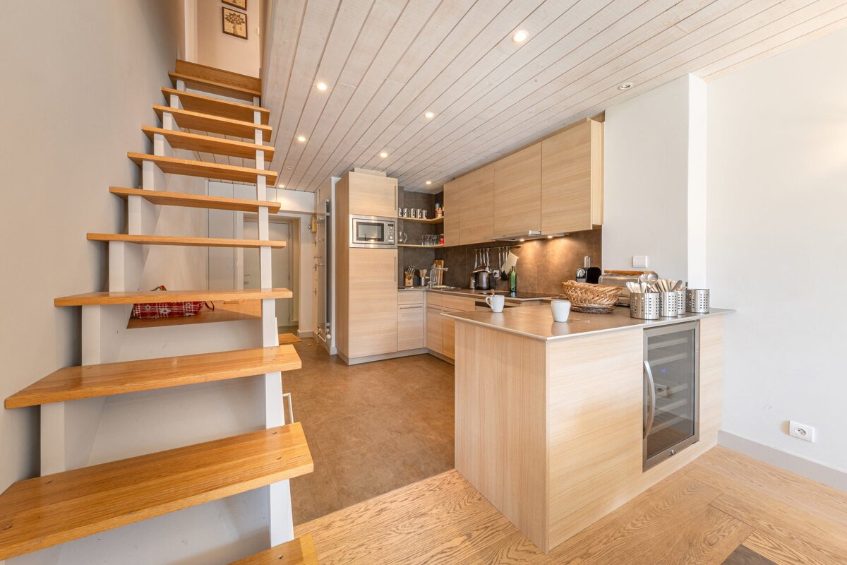 Modern compact apartment featuring open wooden stairs next to a light wood kitchen with an island.