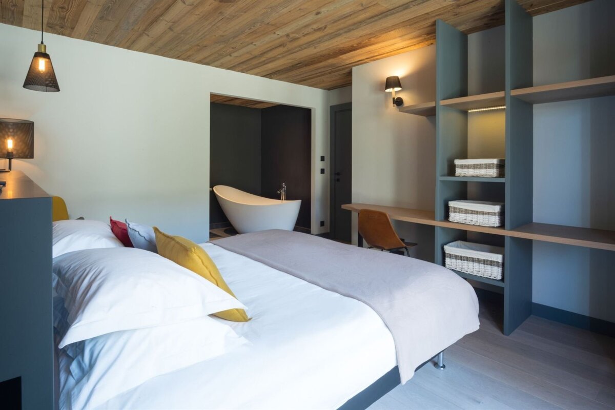Modern bedroom with a white bed, wooden ceiling, and a freestanding bathtub in an adjacent alcove. Features built-in shelves and a desk.