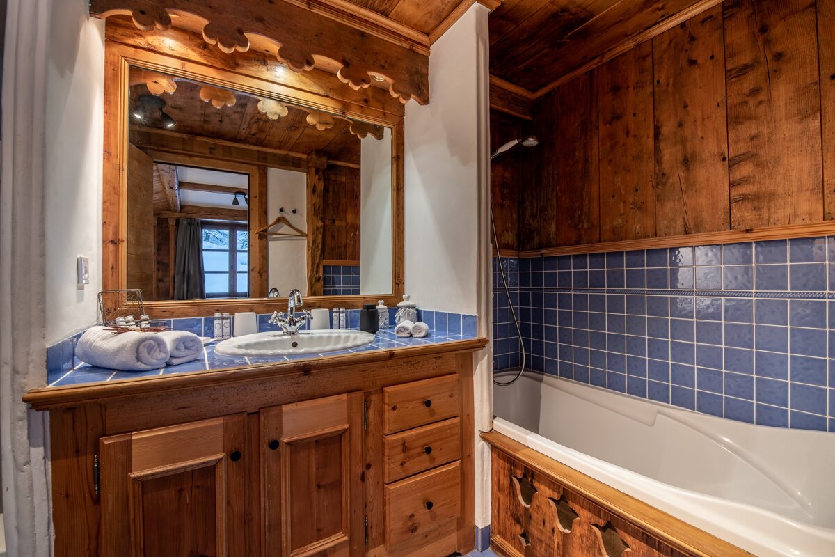A rustic cabin-style bathroom featuring a wooden vanity, a large mirror, and a white bathtub lined with blue tiles.