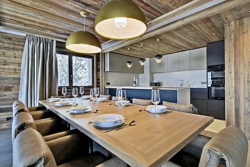 Chic chalet-style dining room with a long wooden table set, gold pendant lights, and a modern open-plan kitchen.
