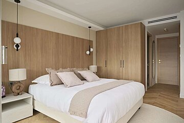 A modern, neutral bedroom features a white bed, wood paneling, integrated wardrobes, and pendant lights. Light wood flooring and subtle decor create a serene ambiance.
