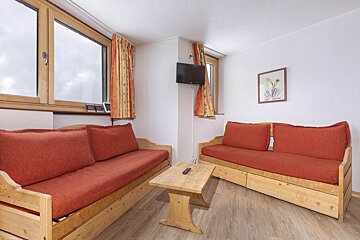 A cozy living area with two wooden daybeds with orange cushions, a wooden coffee table, wall-mounted TV, and windows with orange curtains.