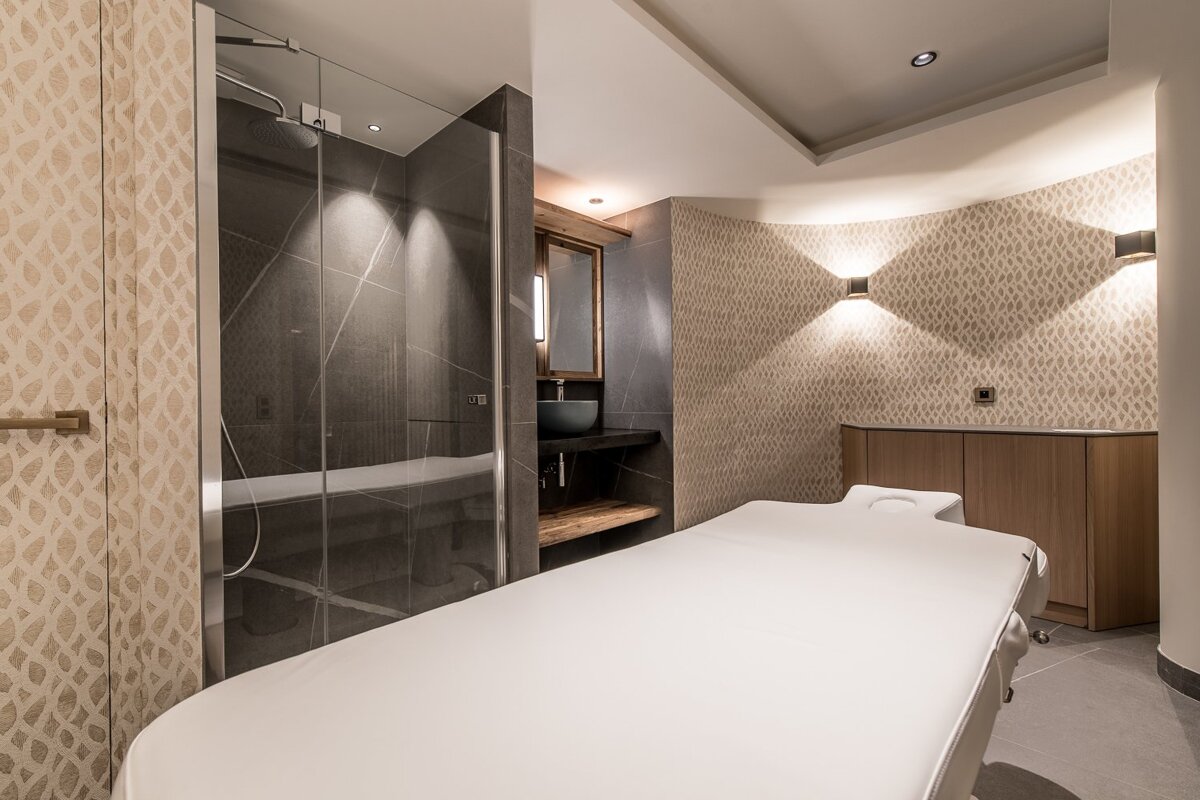 Modern spa room with a white massage table, textured wallpaper, warm accent lighting, and a glass-enclosed walk-in shower.