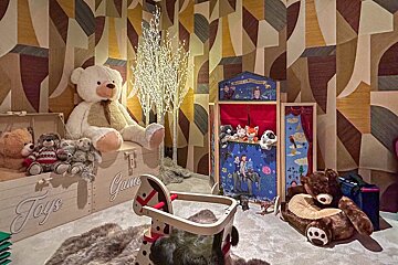 A charming playroom with a giant teddy bear, puppet theater, rocking horse, and various toys, all against a vibrant abstract wallpaper.