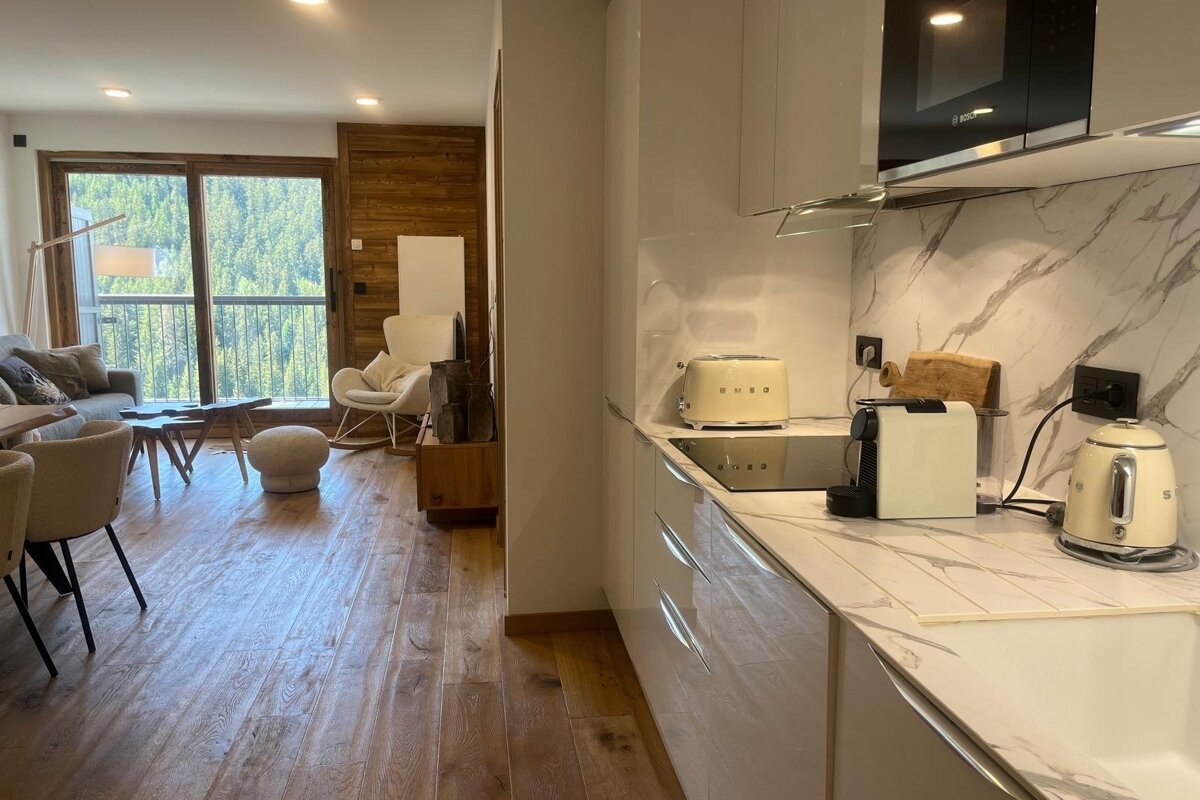A sleek, modern kitchen area adjacent to a cozy living room with wooden floors and balcony views of lush green trees.