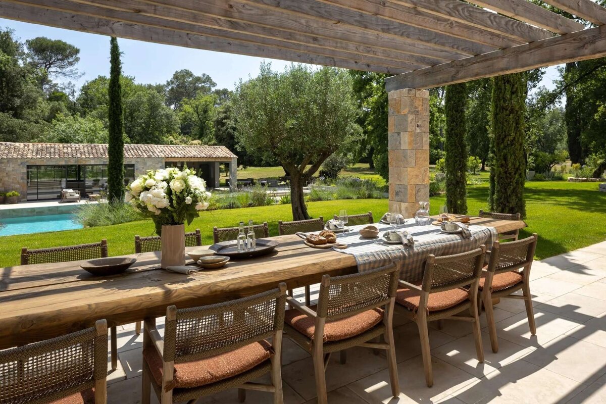 Elegant outdoor dining under a wooden pergola, with a long table set for a meal, overlooking a lush garden, pool, and stone villa.