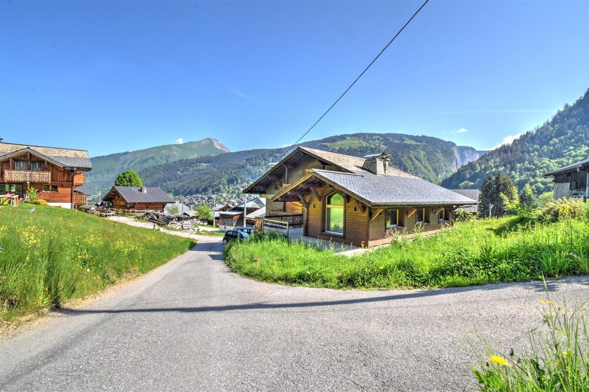 A scenic mountain village with wooden chalets on green hills under a clear blue sky. A winding road leads past houses and lush mountains.