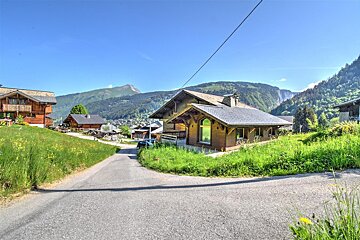 A scenic mountain village with wooden chalets on green hills under a clear blue sky. A winding road leads past houses and lush mountains.