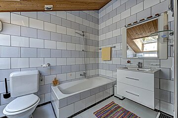 A tiled bathroom features a toilet, bathtub, and vanity under a wooden ceiling. A mirror reflects a skylight and the striped white/grey walls.