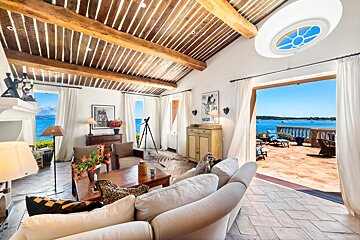 A bright, rustic-chic living room with wood-beam ceilings opening onto a sunny terrace overlooking the ocean.