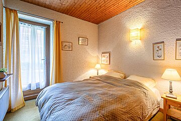 Cozy bedroom with a wooden ceiling, textured walls, a double bed, warm lamps, and yellow curtains by a glass door.
