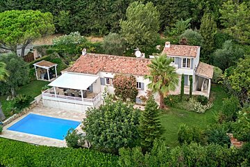 Aerial view: A white house with a red roof, swimming pool, and shaded terrace, surrounded by lush green trees and gardens.