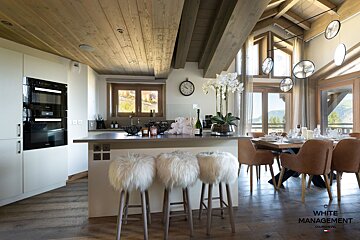 Modern alpine chalet kitchen and dining area featuring cozy white fur barstools and wooden ceilings.