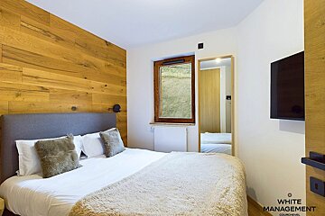 A cozy bedroom features a wooden accent wall, white bed with furry throw, window looking to grass, full-length mirror, and wall-mounted TV.