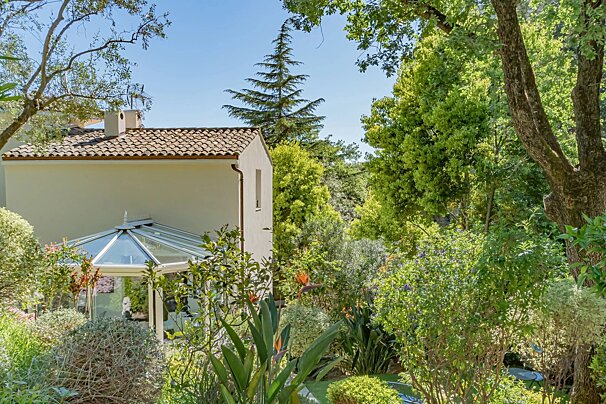 A beige house with a glass conservatory nestled in a lush green garden surrounded by dense trees.