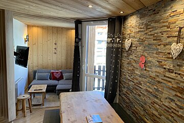 A cozy chalet living space featuring wood paneling, a stone accent wall, a gray sofa, and a wooden dining table by a window with patterned curtains.