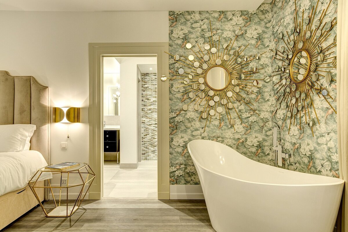 A luxurious room featuring a bed, a freestanding tub, and ornate sunburst mirrors on floral wallpaper, with a bathroom visible through a doorway.