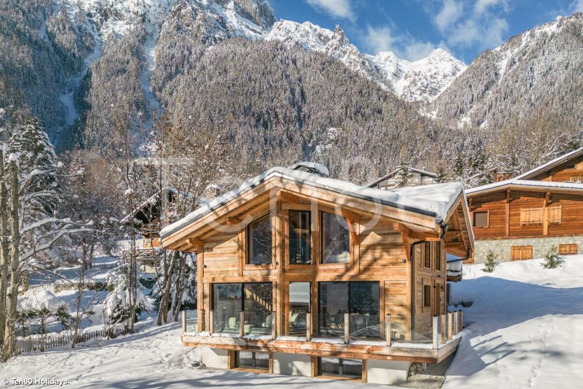 A cozy wooden chalet stands in deep snow, surrounded by a winter wonderland of snow-covered trees and towering mountains under a blue sky.