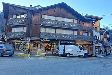 A rustic wooden chalet-style building with shops (Fromagerie, La Loge), ornate balconies, and two vans parked on a street.