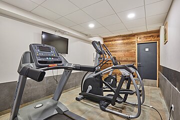 A modern gym features a treadmill, elliptical, wall-mounted TV, rustic wooden accent wall, and tiled floors.