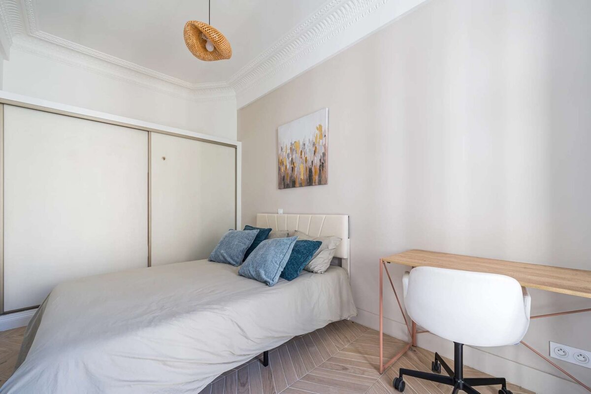 A minimalist bedroom with a bed, blue pillows, white closet, desk, and chair. Features light walls, a woven pendant light, and abstract art.